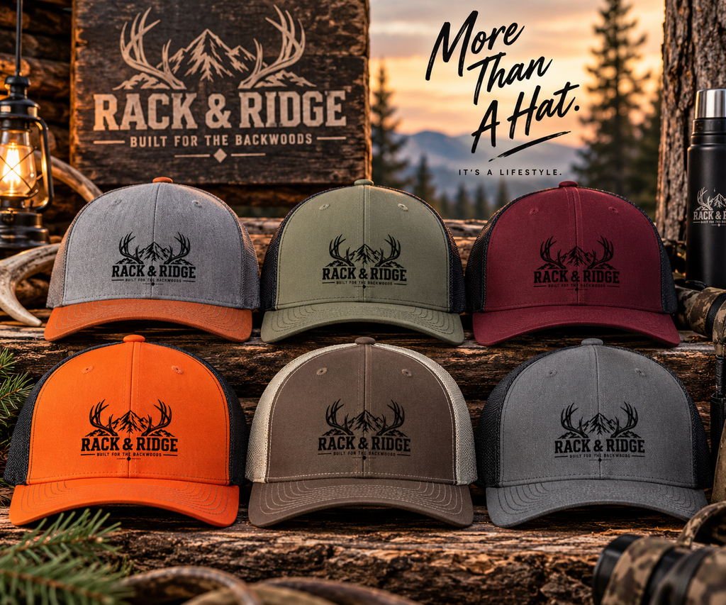 Six Rack & Ridge trucker hats in assorted colors are arranged on rustic logs with outdoor-themed props, including a branded wooden sign, a lantern, trees, and a black insulated tumbler. The hats feature black mesh back panels and a black embroidered antler logo with the text 'RACK & RIDGE' and 'BUILT FOR THE BACKWOODS.